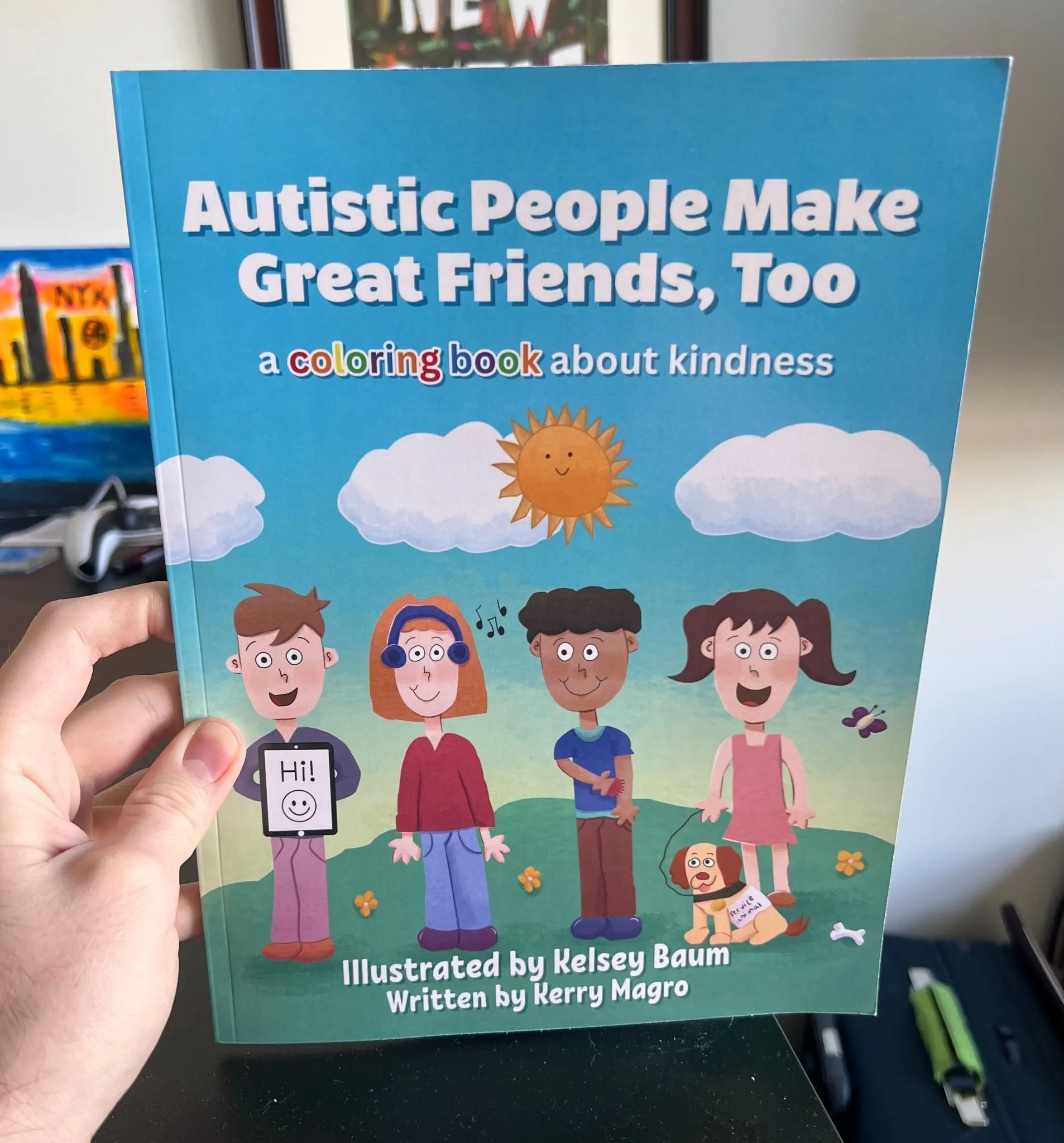 A hand holds up the cover of "Autistic People Make Great Friends, Too: A Coloring Book About Kindness," showing four illustrated children and a service dog under a sunny sky, illustrated by Kelsey Baum and written by Kerry Magro.