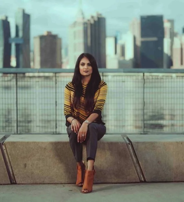 Dr. Sneha Gazi, founder of Sneha Physical Therapy, sitting on a concrete ledge with the Manhattan skyline behind her.